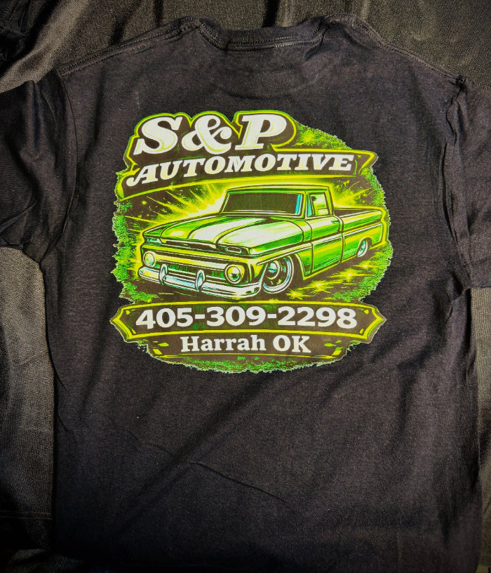 A dark charcoal gray t-shirt laid flat, featuring a large graphic print on the back with a vintage green and white pickup truck and the text 'S&P Automotive 405-309-2298 Harrah OK'.