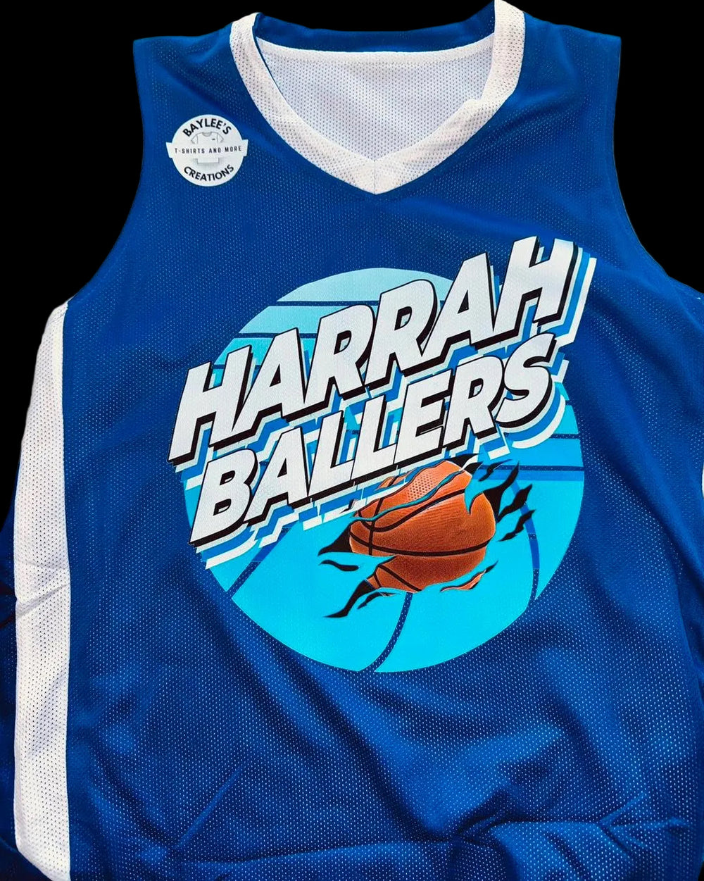 A blue mesh basketball jersey laid flat on a light surface, featuring a large circular graphic with the words “HARRAH BALLERS” in white outlined lettering over a light blue background, plus an orange basketball illustration. A small round patch near the upper left chest reads “Wellesley High School Champions.