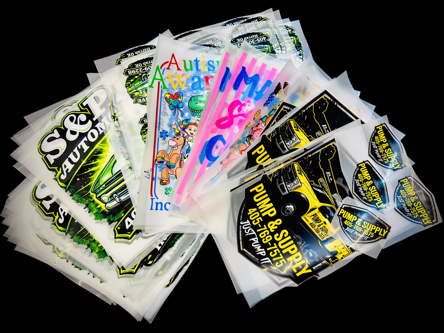 A pile of DTF Prints, including designs with 'S&P CAUTION', 'Auto Parts Warehouse', 'PUMP & SUPPLY', and cartoon graphics, arranged on a black textured background.