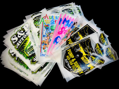 A pile of DTF Prints, including designs with 'S&P CAUTION', 'Auto Parts Warehouse', 'PUMP & SUPPLY', and cartoon graphics, arranged on a black textured background.