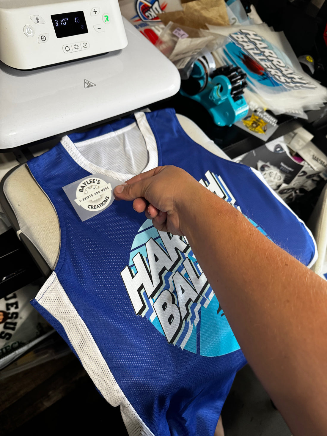 A blue and white basketball jersey with a custom graphic print, being held by a hand near a heat press machine.
