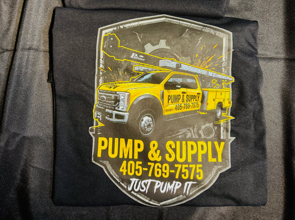 A folded black t-shirt laid flat on a dark fabric background. The top portion shows a small bright green 'S&P' logo on the chest, while the lower portion displays a large shield-style graphic featuring a yellow pump truck and the text 'PUMP & SUPPLY 405-769-7575 JUST PUMP IT'.